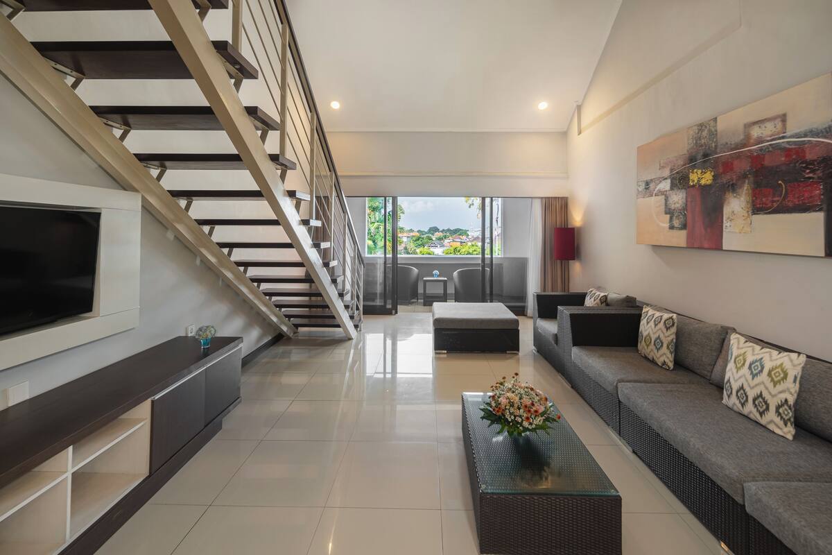 A modern living room with a staircase, a flat-screen TV, a sofa set, and a coffee table with flowers.