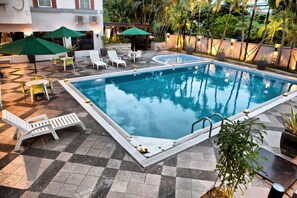 A rectangular pool with a tiled deck, surrounded by lounge chairs and umbrellas, and a small hot tub.