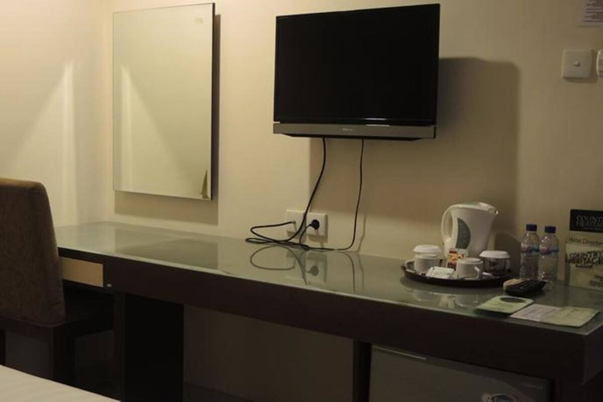 A hotel room with a desk, a television mounted on the wall, a chair, a coffee maker, and a tray with cups and a remote control.