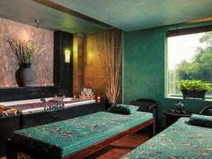 A spa room with a large bathtub, two beds with patterned bedding, a small table with candles, and a window with a view of greenery.