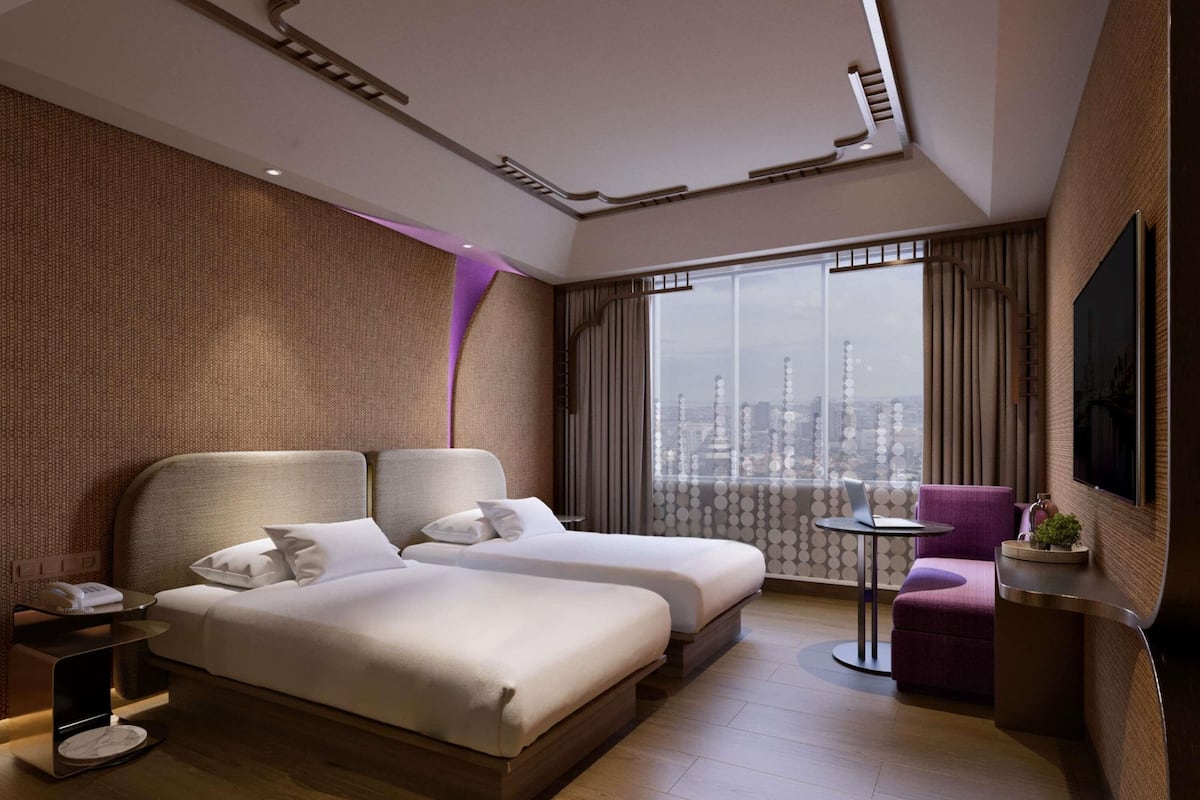 Una habitación de hotel moderna con vista a la ciudad, dos camas, una mesita, una silla y un televisor.