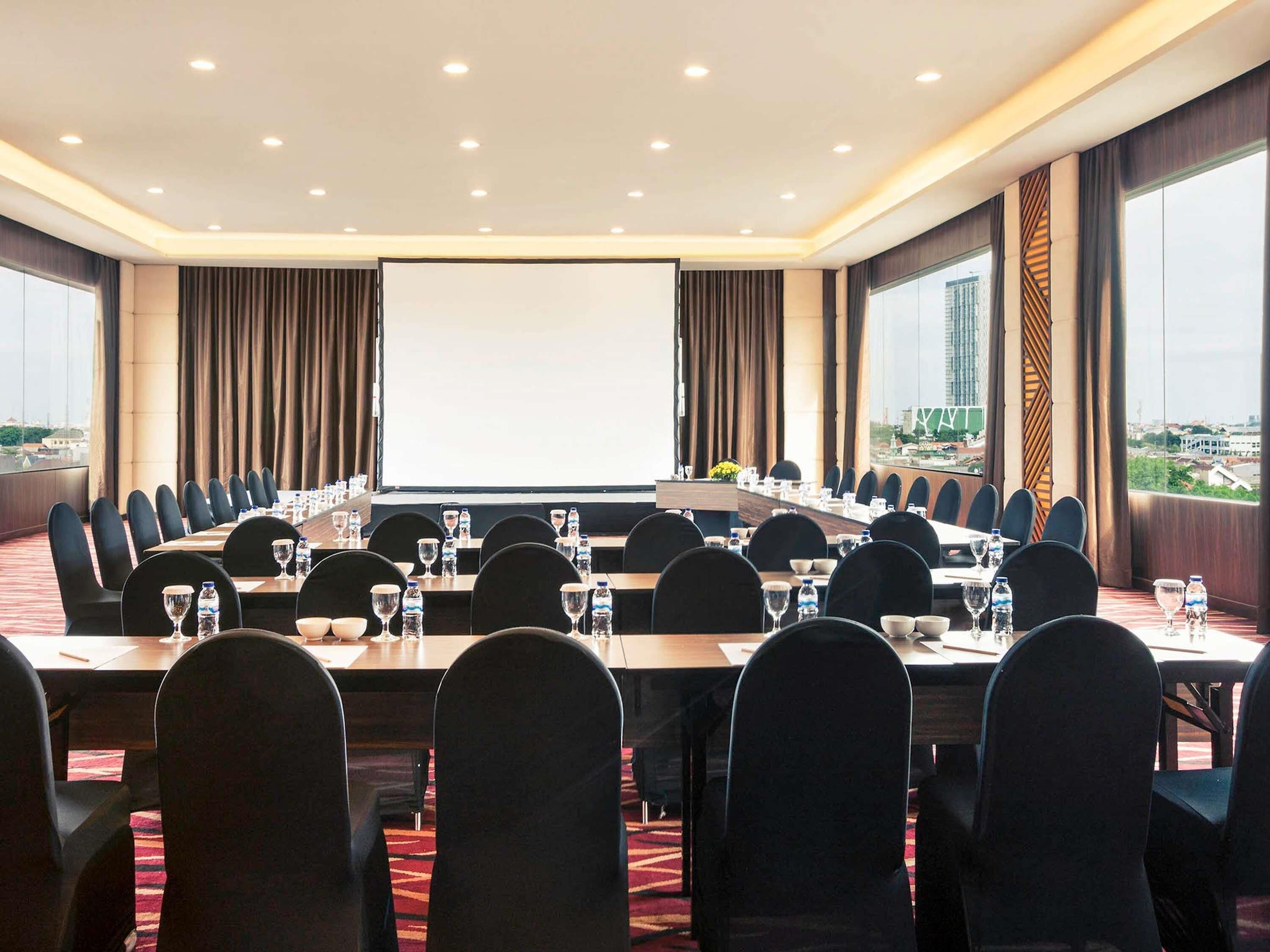 Un salón de conferencias con una pantalla grande, varias mesas y sillas dispuestas para reuniones.