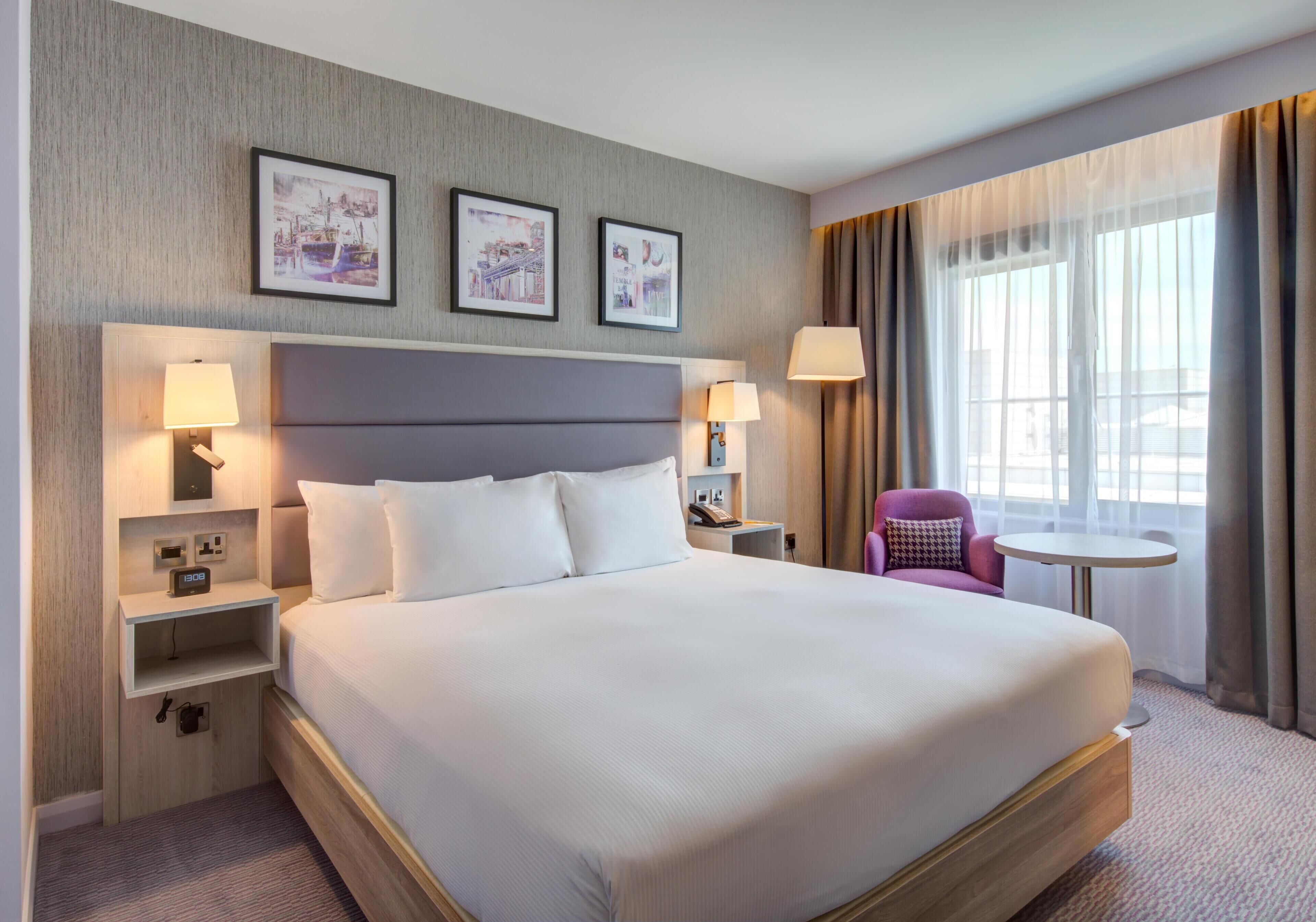 Habitación de hotel con una cama grande, dos mesitas de noche con lámparas, una mesa redonda pequeña, una silla morada y cuadros enmarcados en la pared.