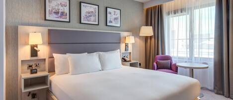 Habitación de hotel con una cama grande, dos mesitas de noche con lámparas, una mesa redonda pequeña, una silla morada y cuadros enmarcados en la pared.