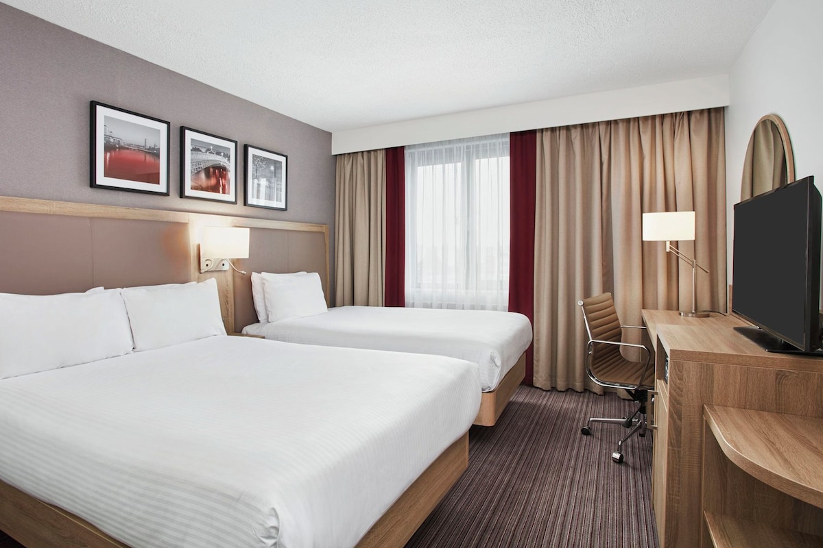 A hotel room with two beds, a desk with a television, a chair, and a window with curtains.