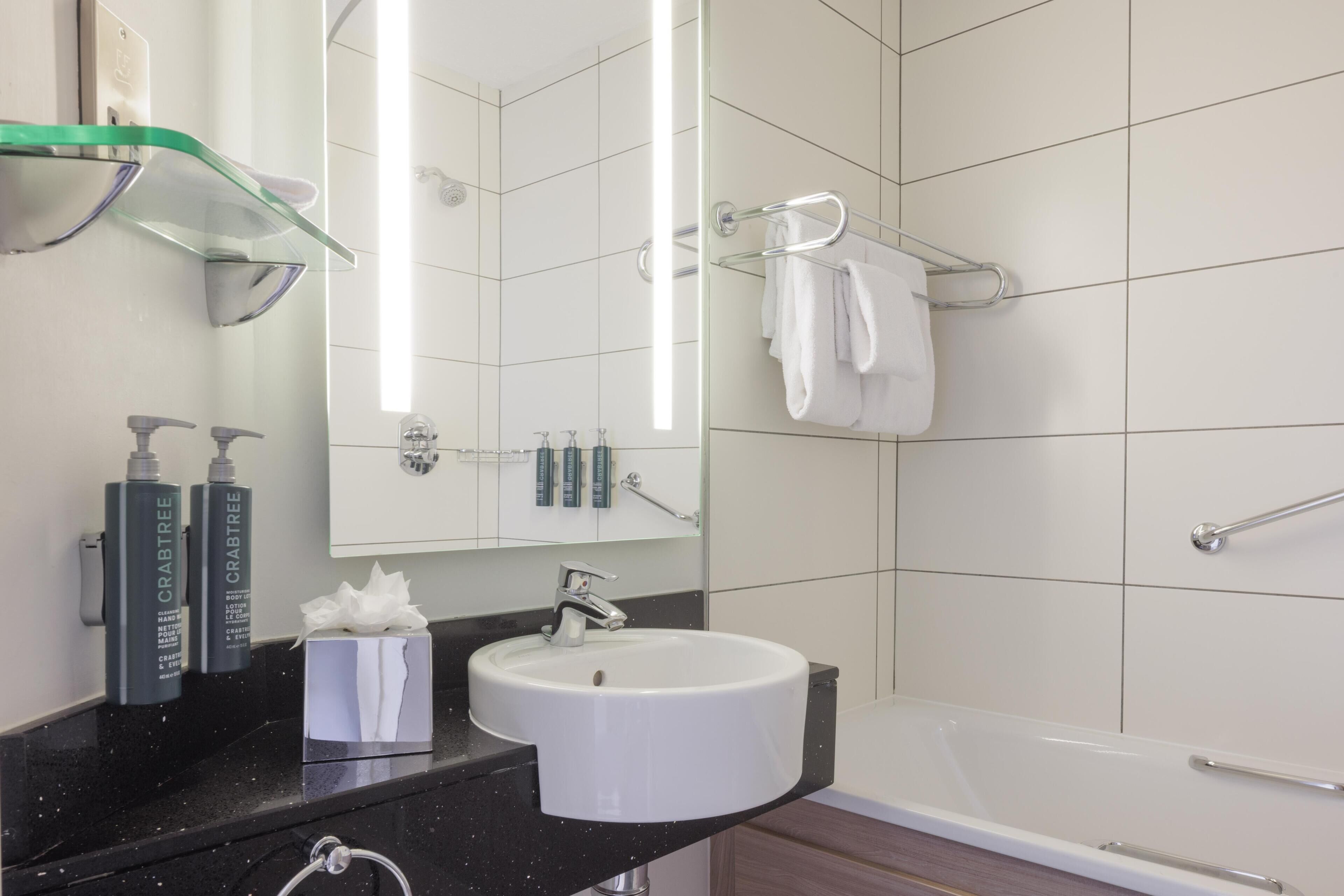 A modern bathroom with a white sink, mirror, and bathtub. There are toiletries on the counter and a towel rack on the wall.