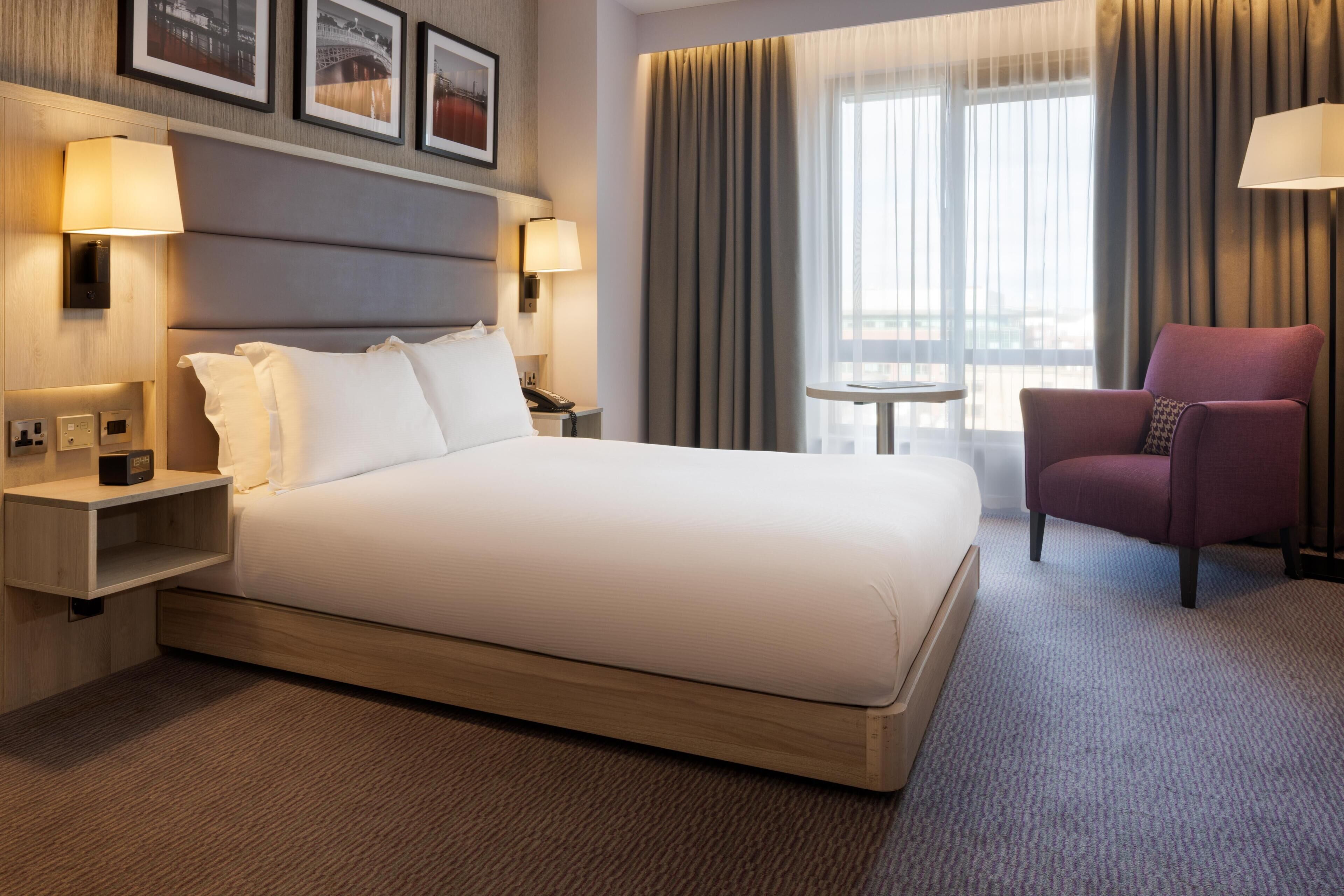 Habitación de hotel con una cama grande, lámparas de noche, una mesita, una silla y vistas a la ciudad desde la ventana.