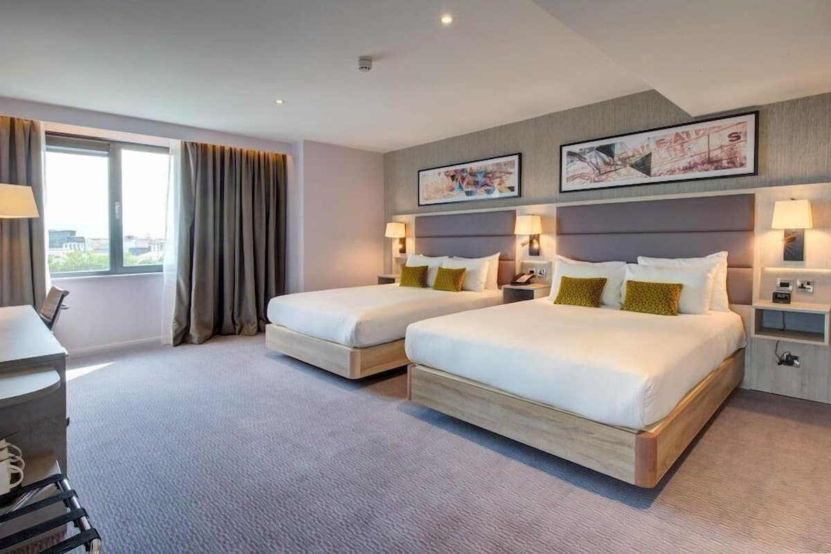 A hotel room with two beds, a desk, and a large window with curtains.