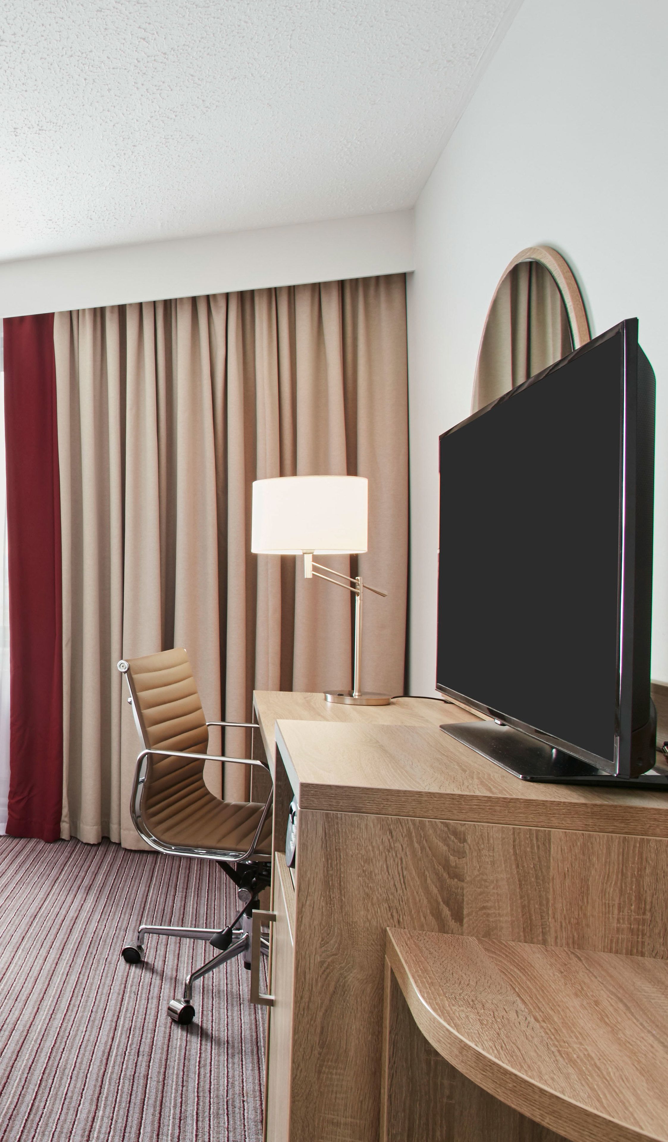 A hotel room with a wooden desk, a television, a chair, a lamp, and a large window with curtains.
