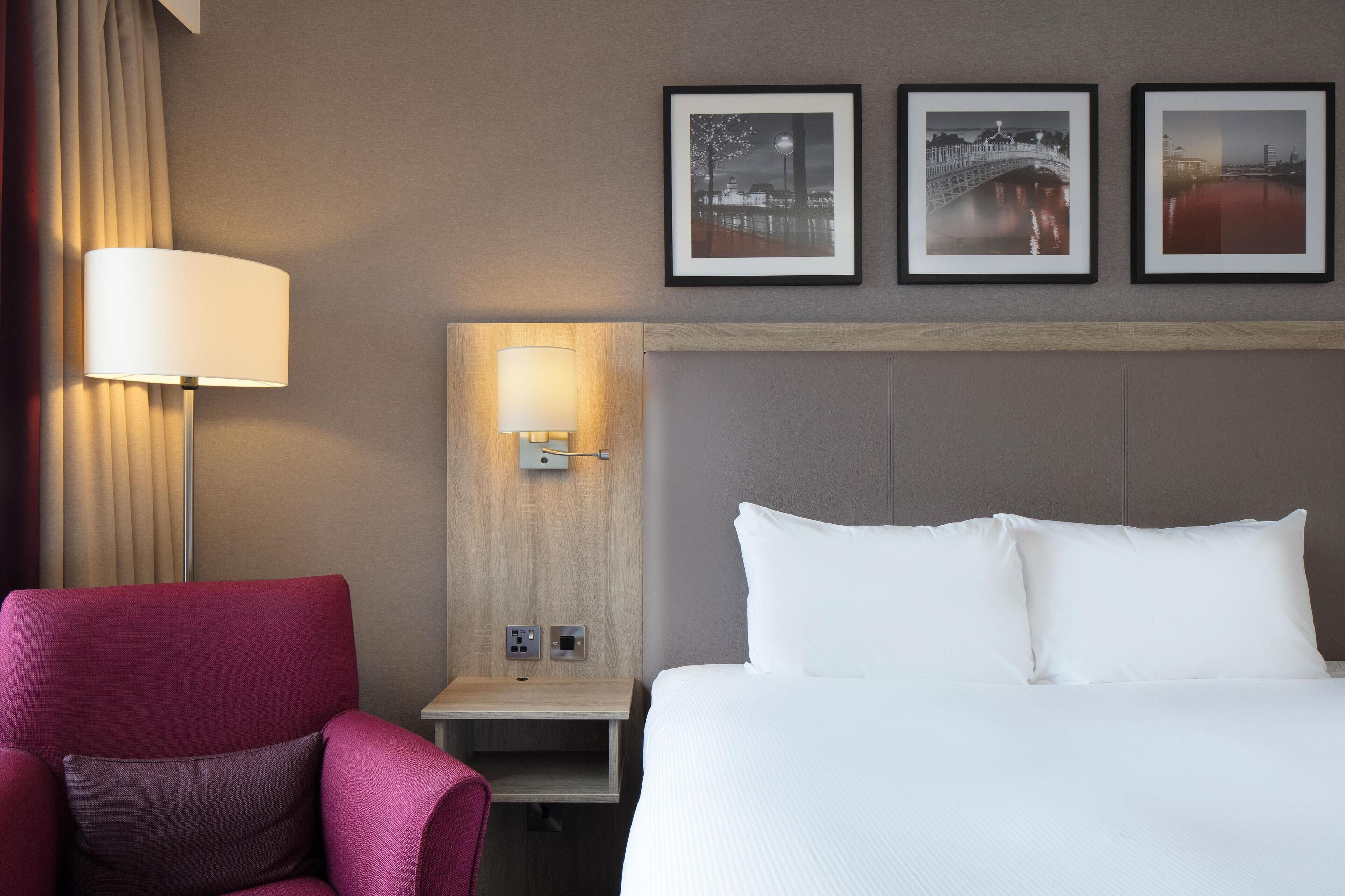 Una habitació d'hotel amb un llit, una tauleta de nit, una làmpada, una cadira i tres quadres penjats a la paret.