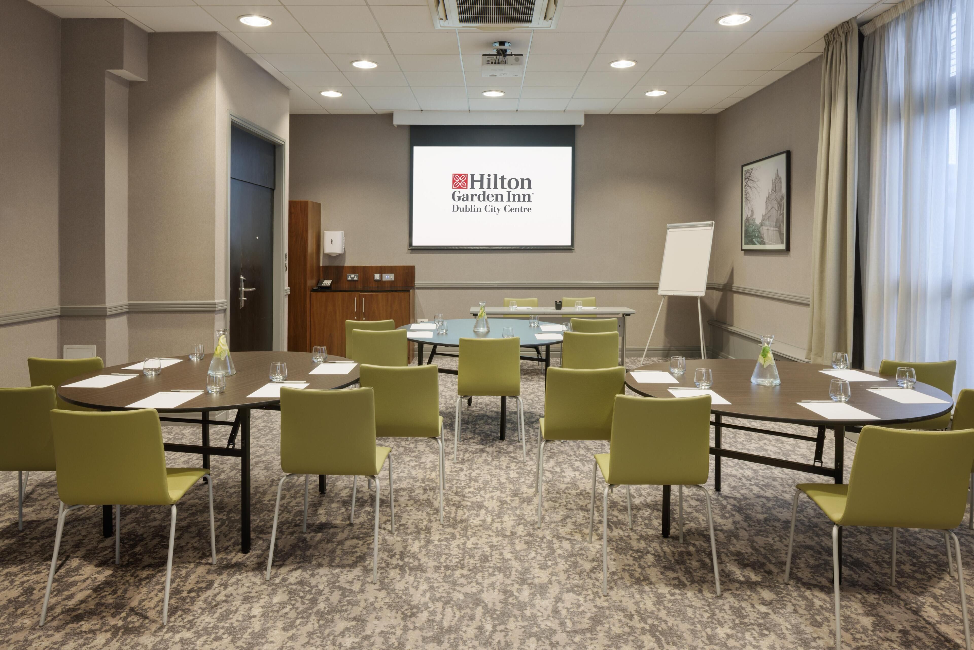 A conference room with a large screen displaying the Hilton Garden Inn logo, green chairs around a round table, and a carpeted floor.