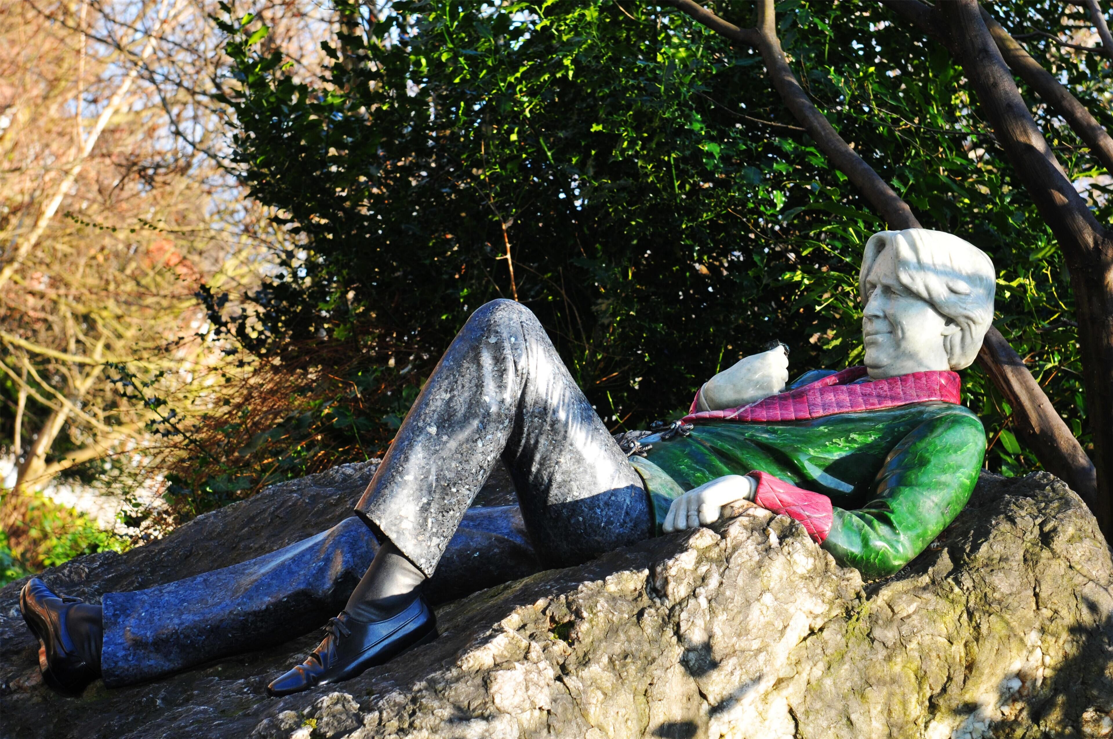 Une statue représentant une personne portant une veste verte et un pantalon bleu, assise sur un rocher.