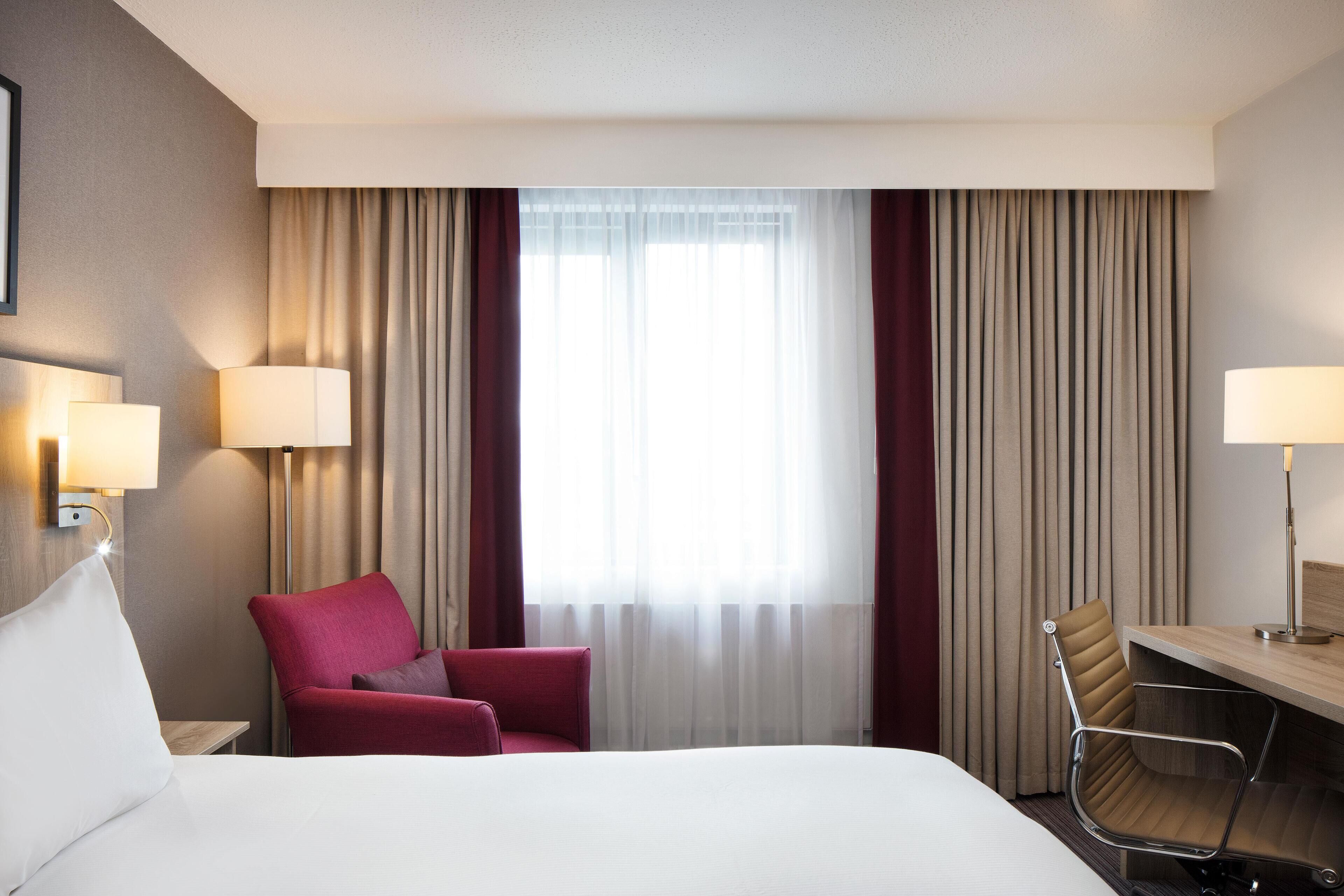 A hotel room with a bed, a desk, a chair, a lamp, and a window with curtains.
