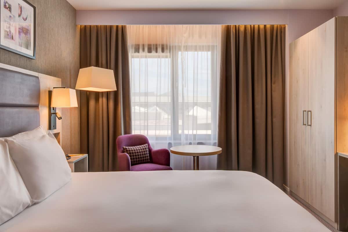 A hotel room with a bed, a purple armchair, a small round table, a lamp, and a large window with curtains.