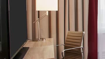 Une chambre d’hôtel équipée d’un bureau, d’une télévision, d’une chaise, d’une lampe et d’une grande fenêtre avec des rideaux.