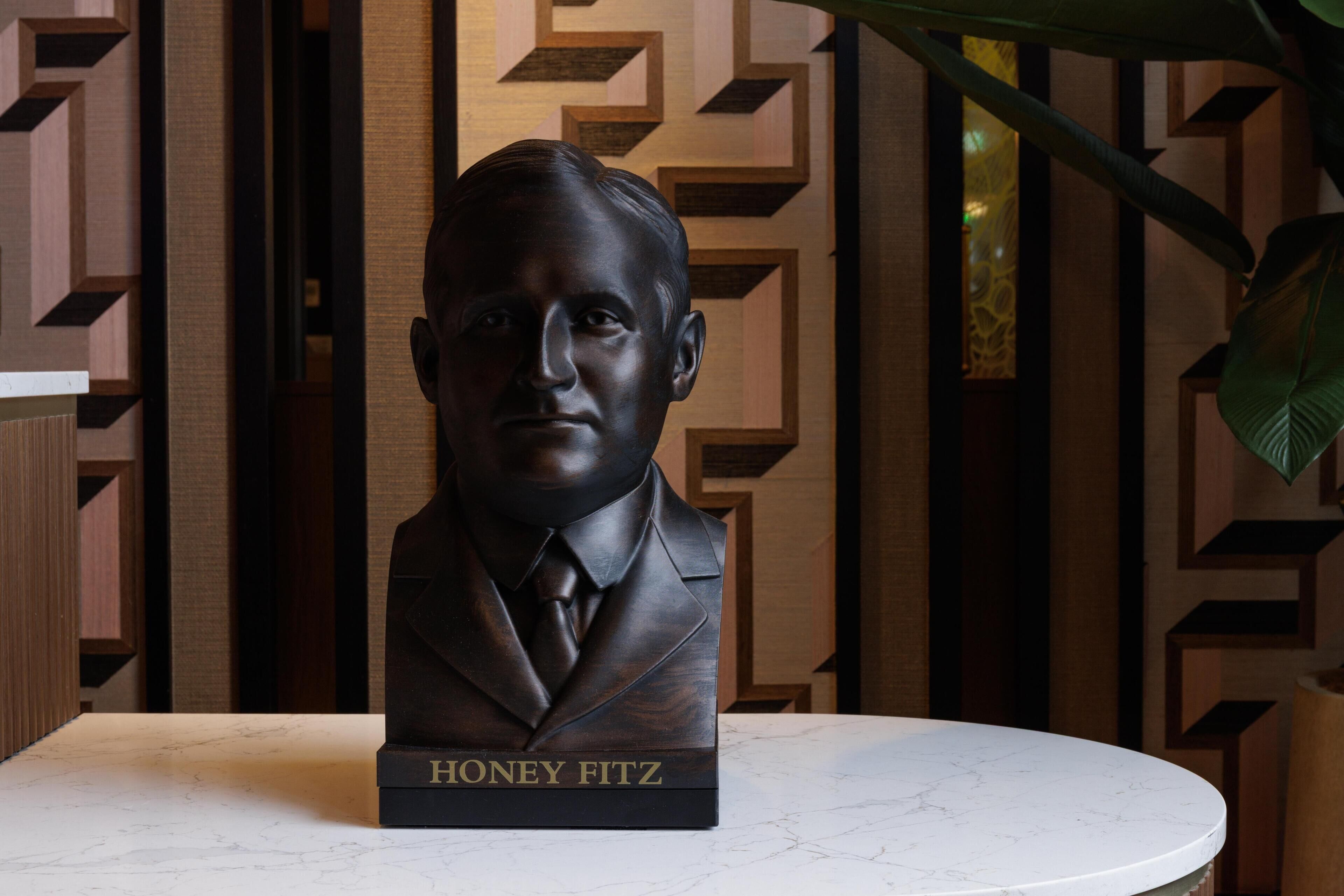 A bust of Honey Fitz on a marble table with a geometric patterned wall in the background.