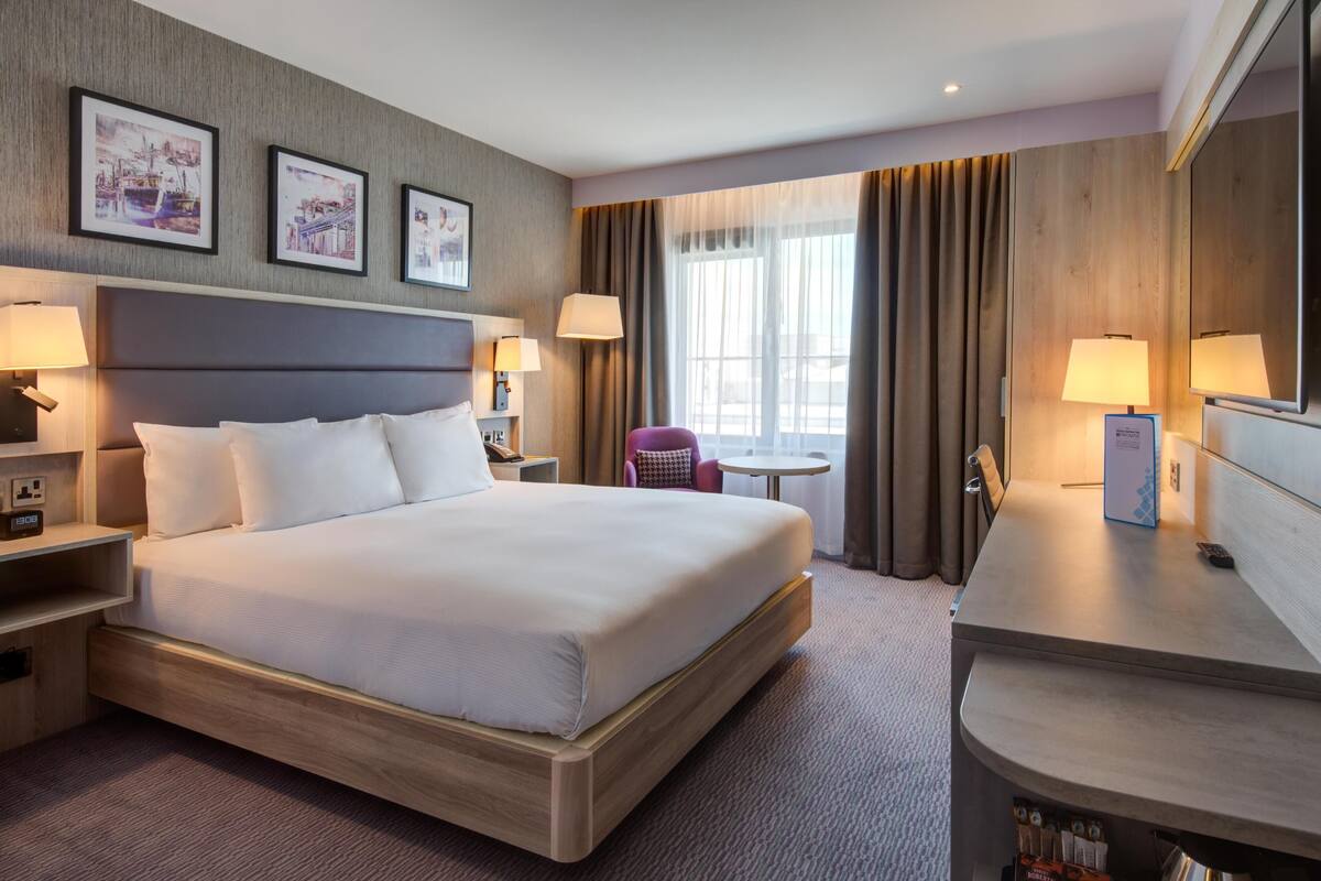 A hotel room with a large bed, bedside tables, a desk, a chair, and framed pictures on the wall.