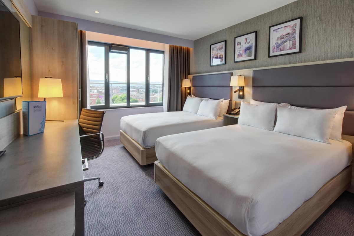 A hotel room with two beds, a desk, a chair, a lamp, and a window with a city view.