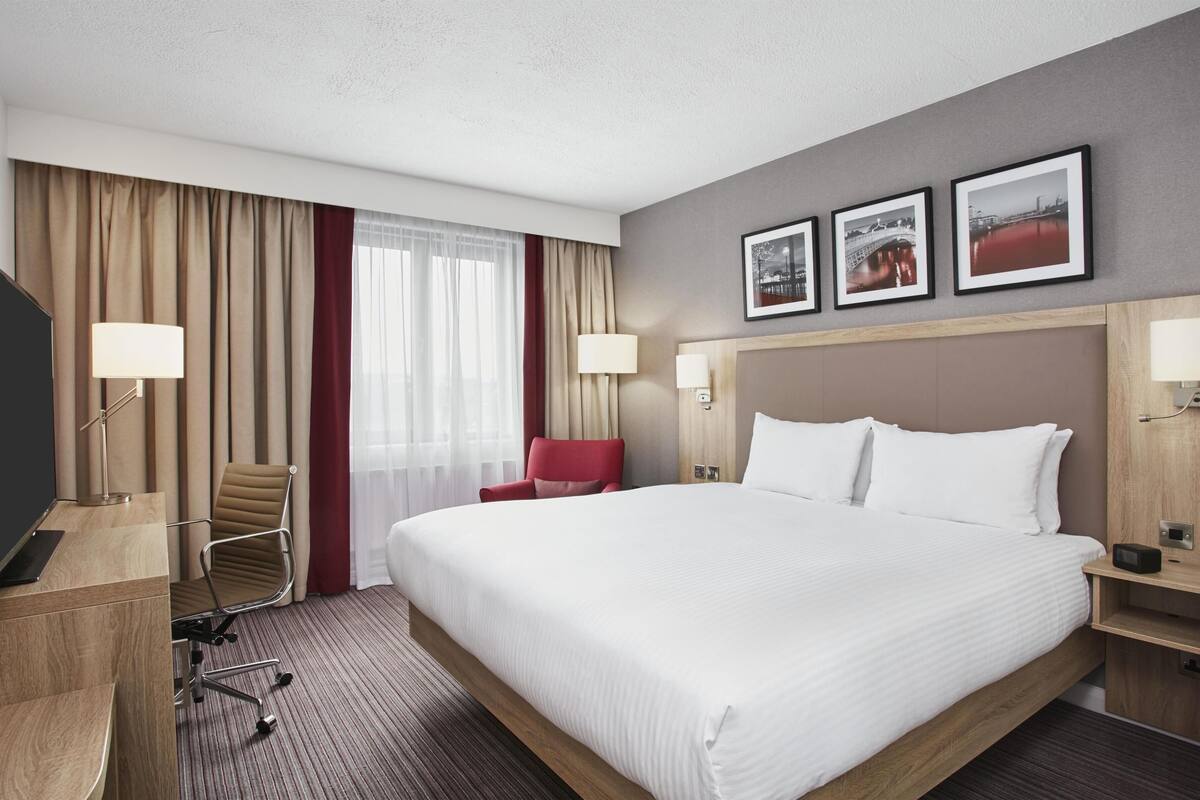 A hotel room with a large bed, a desk, a chair, and two wall-mounted lamps.