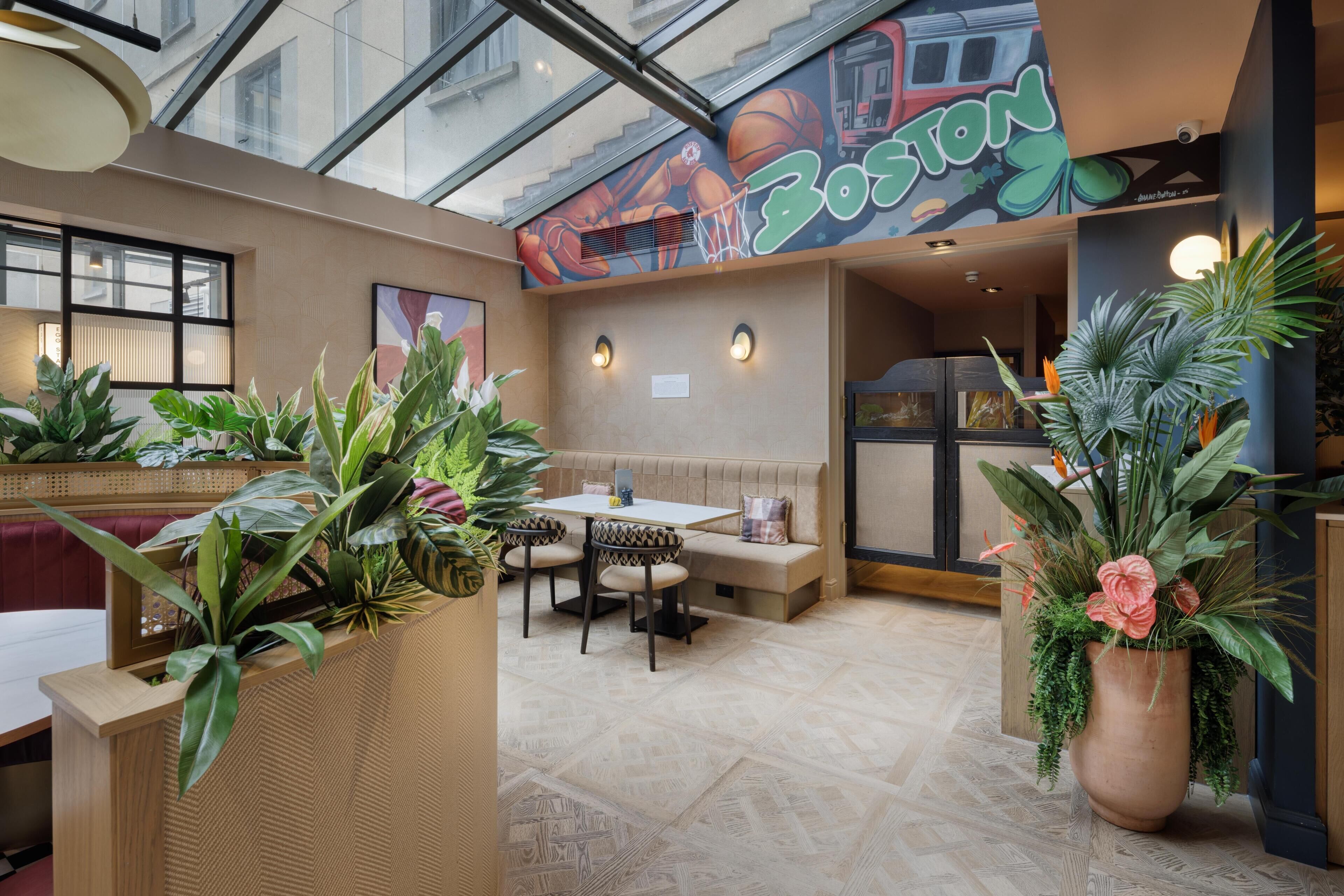 A restaurant with a high ceiling, indoor plants, a seating area with a bench, and a mural on the wall.