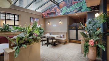 A restaurant with a high ceiling, indoor plants, a seating area with a bench, and a mural on the wall.
