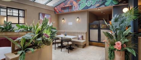 A restaurant with a high ceiling, indoor plants, a seating area with a bench, and a mural on the wall.
