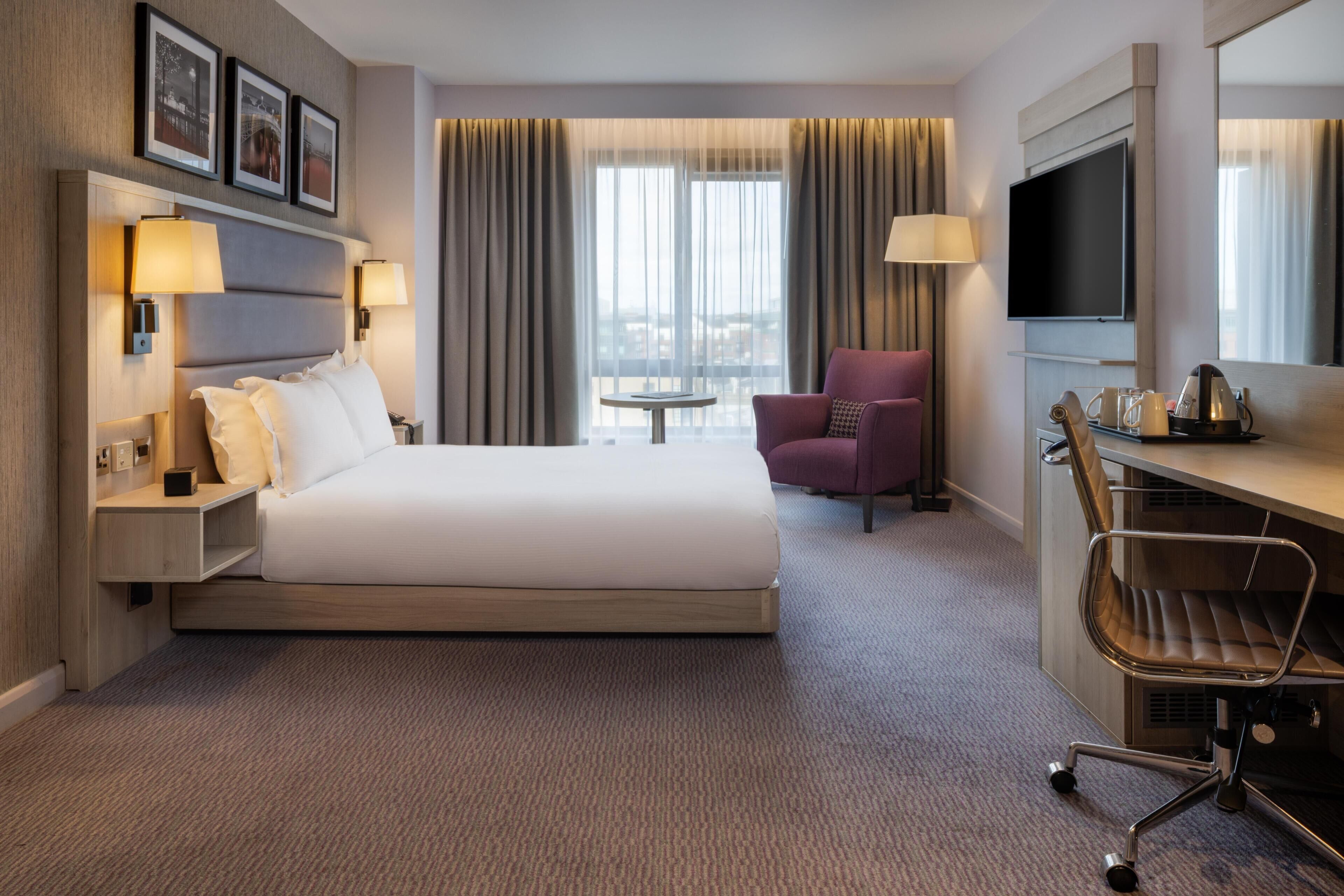 A hotel room with a bed, a desk, a chair, a TV, and a window with curtains.