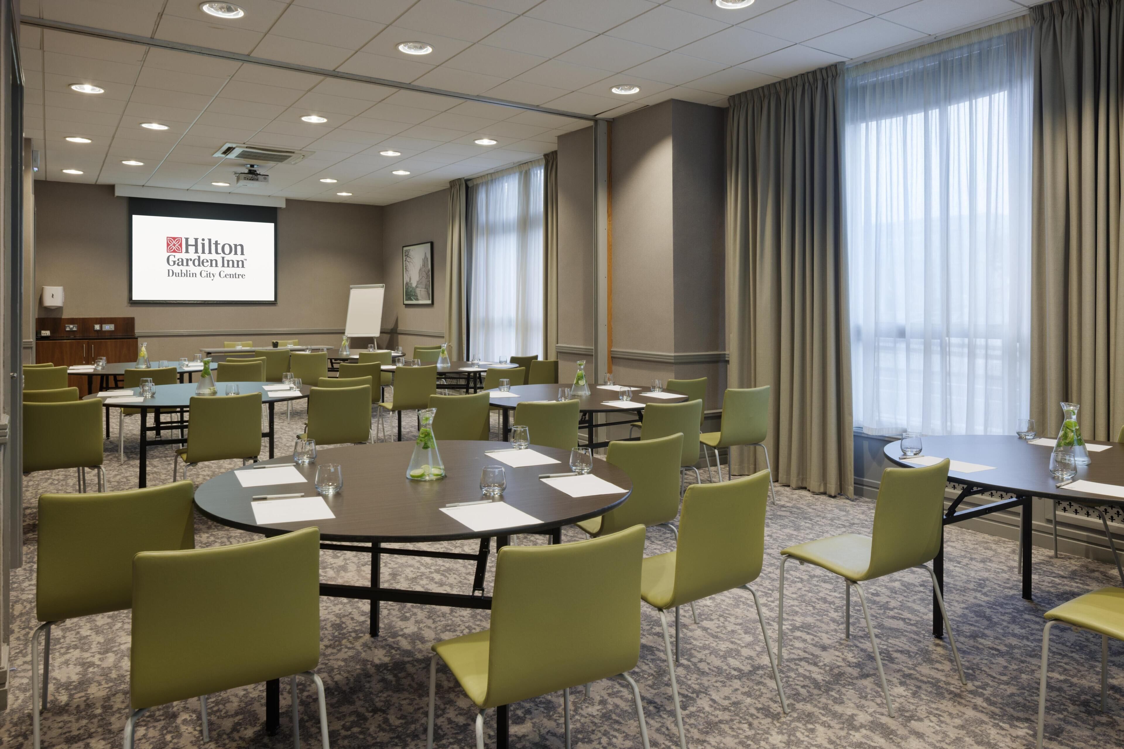 A conference room with multiple tables and chairs, a projector screen displaying Hilton Garden Inn logo, and large windows with curtains.