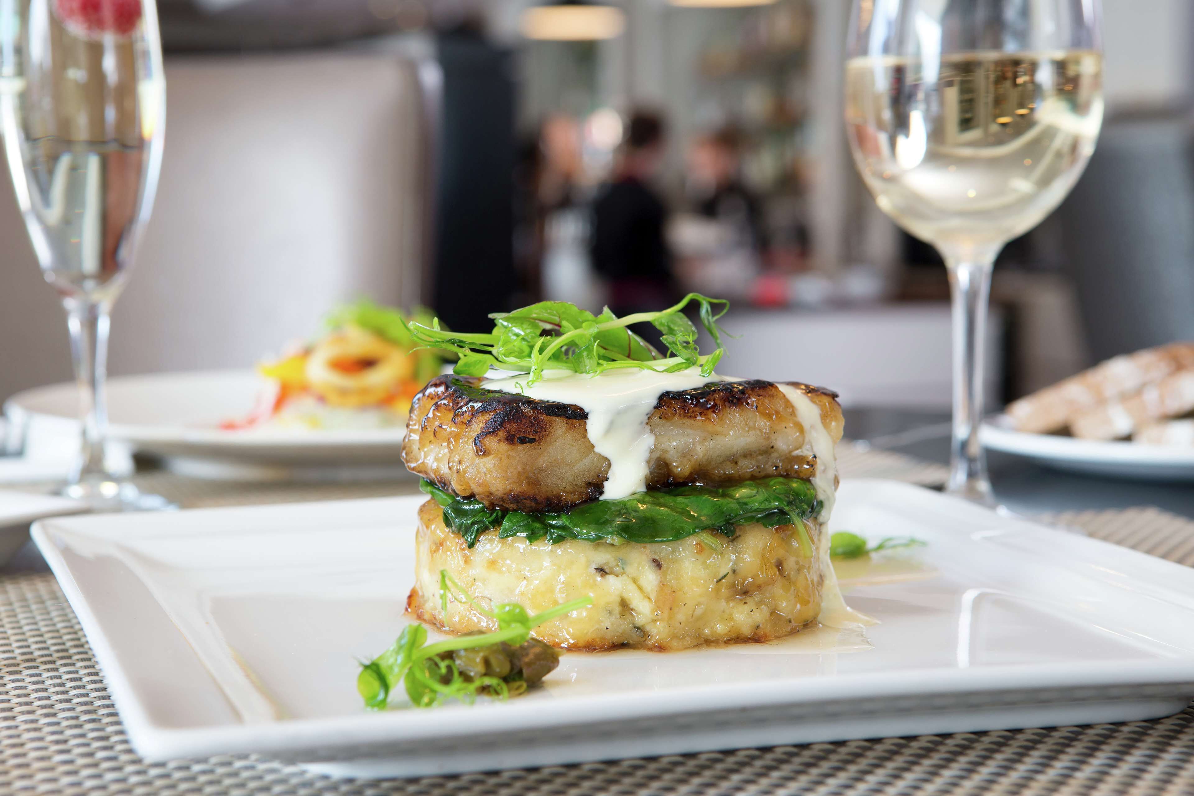 A plated dish with a creamy sauce, greens, and a piece of meat, accompanied by a glass of white wine.