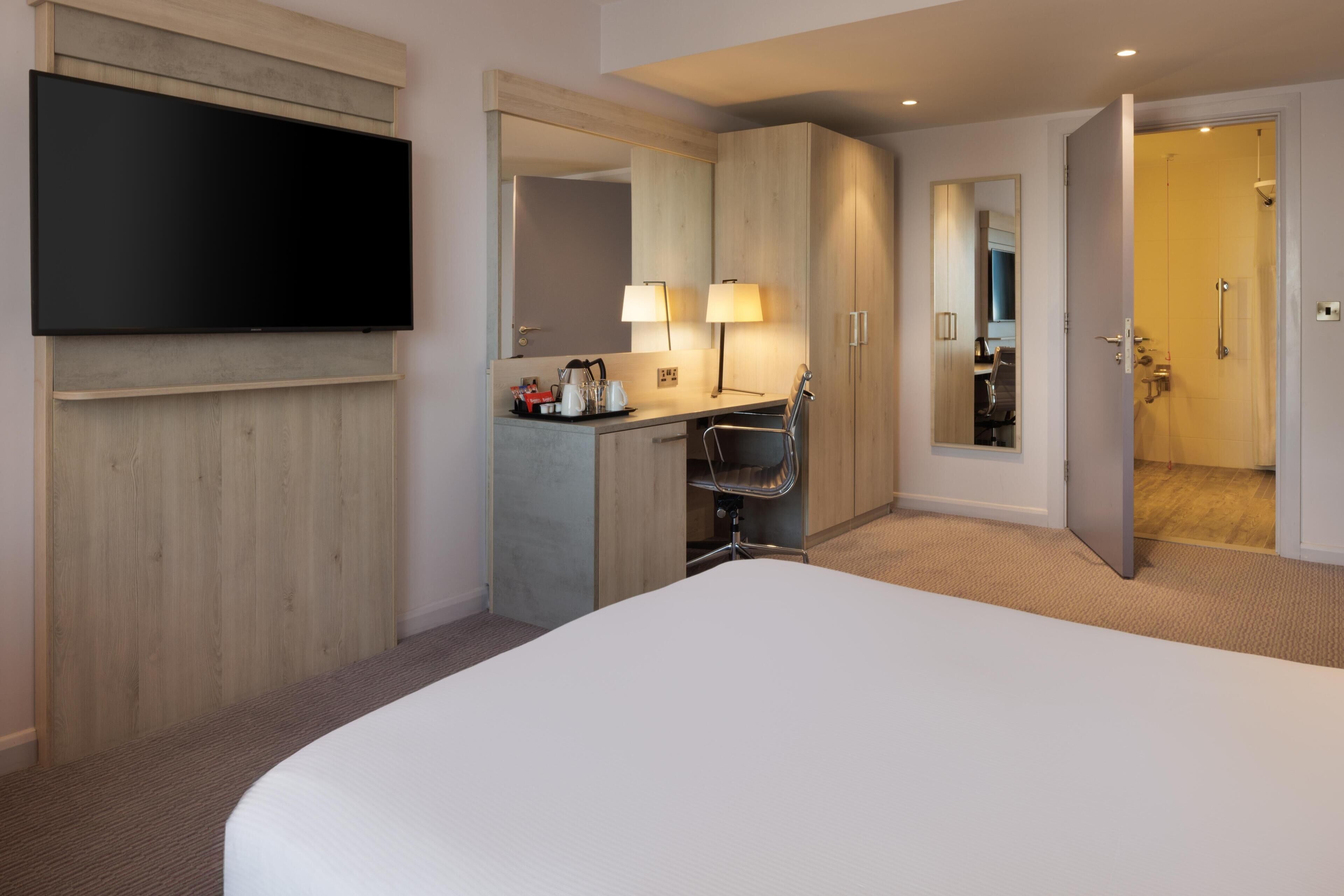 Habitación de hotel con cama, televisión, escritorio con silla, espejo y puerta que da a un baño.