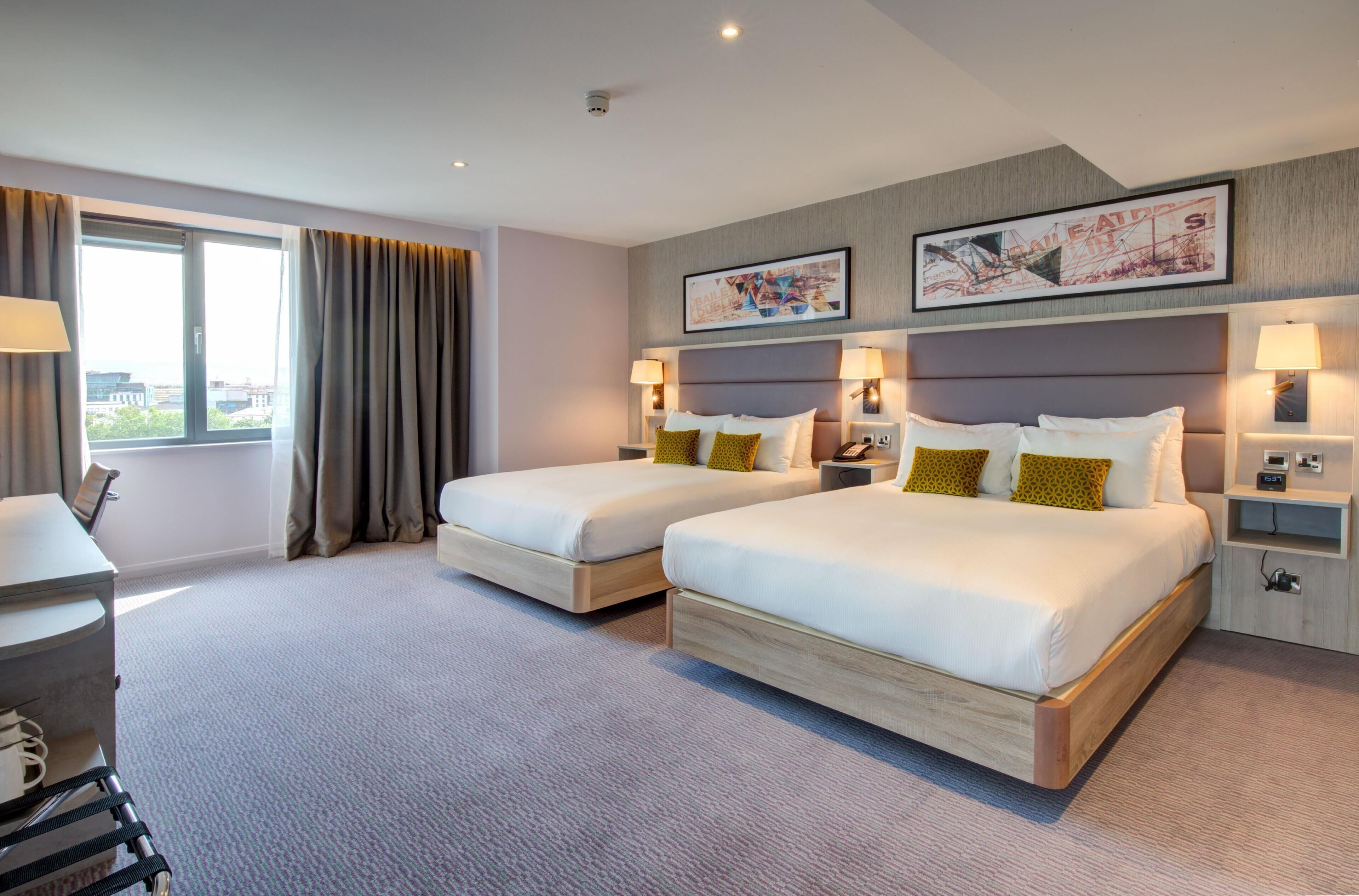 A hotel room with two beds, a desk, a chair, a window with curtains, and two wall-mounted lamps.