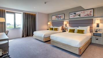 A hotel room with two beds, a desk, a chair, a window with curtains, and two wall-mounted lamps.
