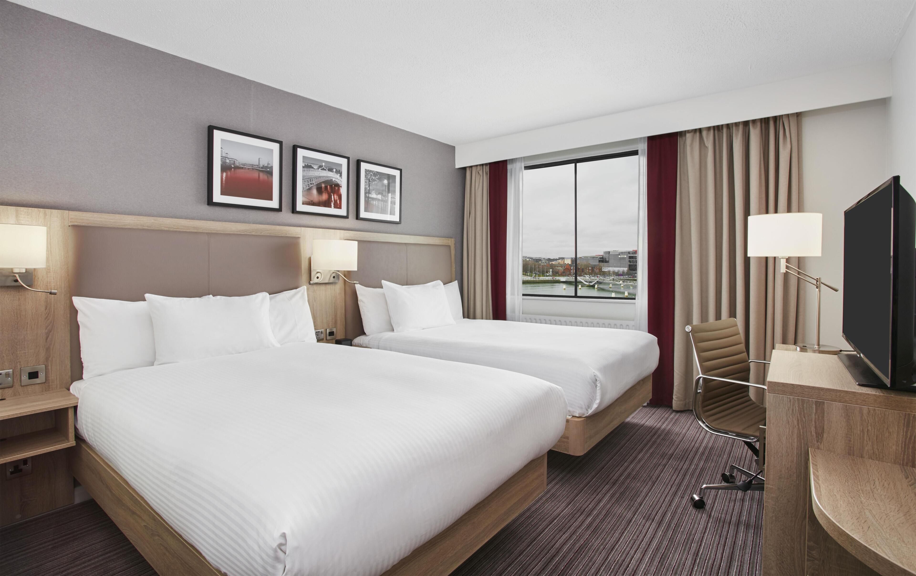 Ein Hotelzimmer mit zwei Betten, einem Fernseher, einem Schreibtisch und einem Fenster mit Blick auf die Stadt.