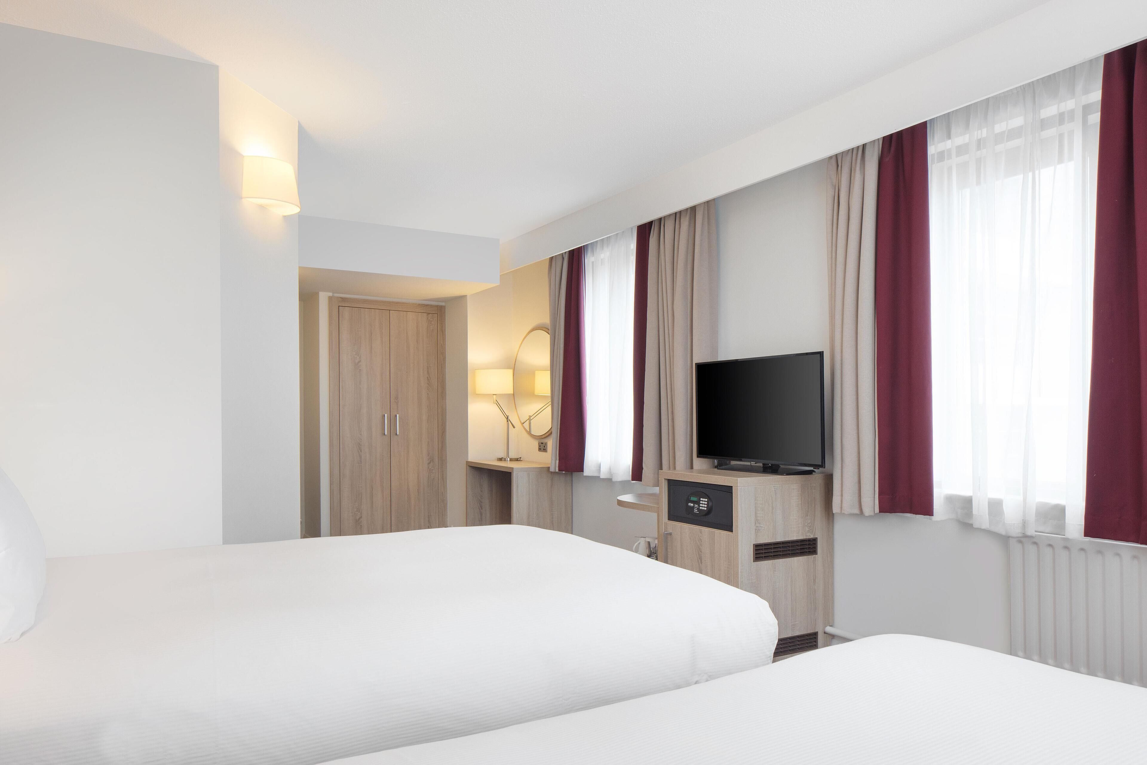 A hotel room with two beds, a TV, a desk with a lamp, and a window with curtains.