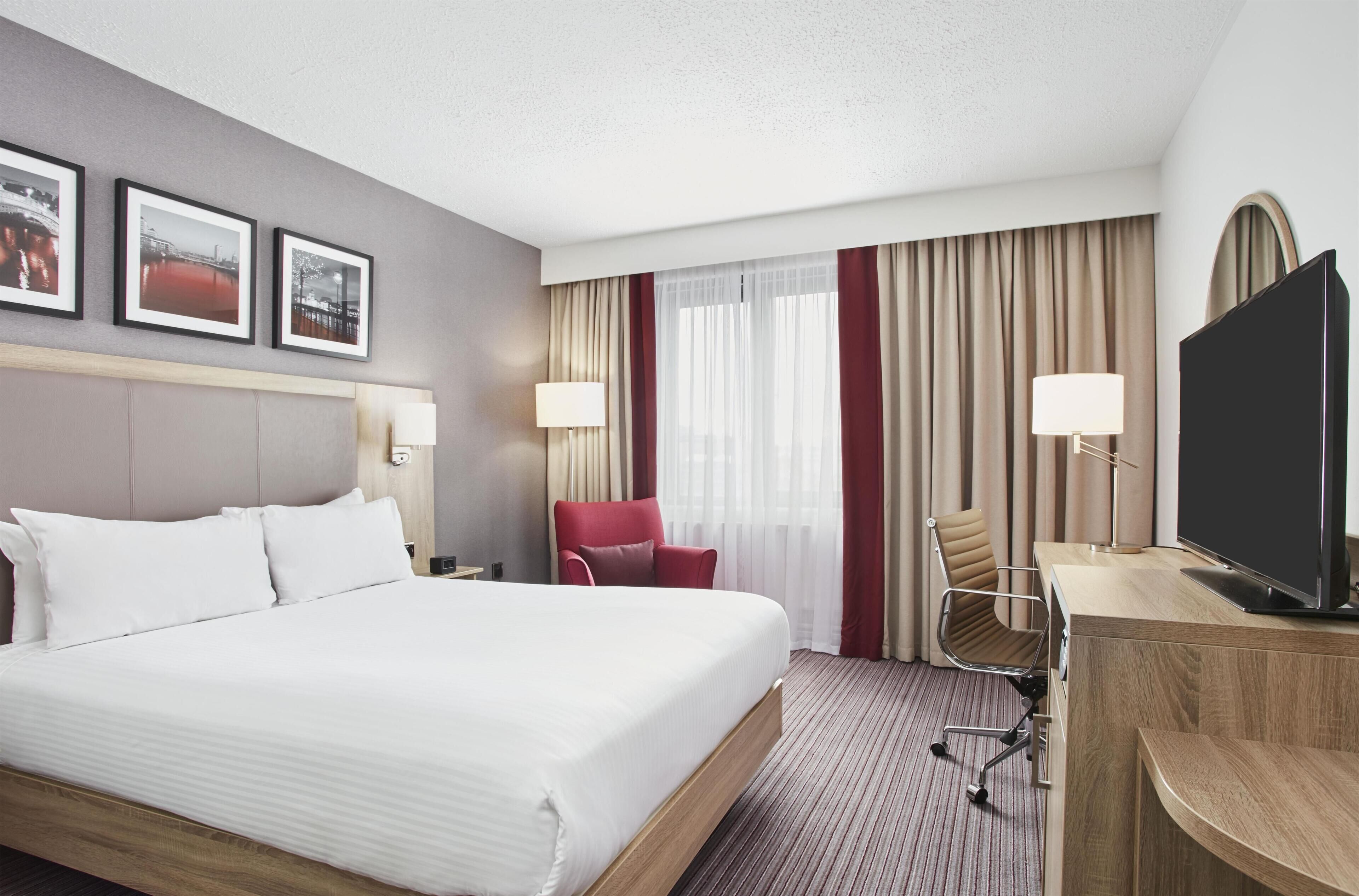 A hotel room with a bed, a desk with a TV, a chair, and a window with curtains.