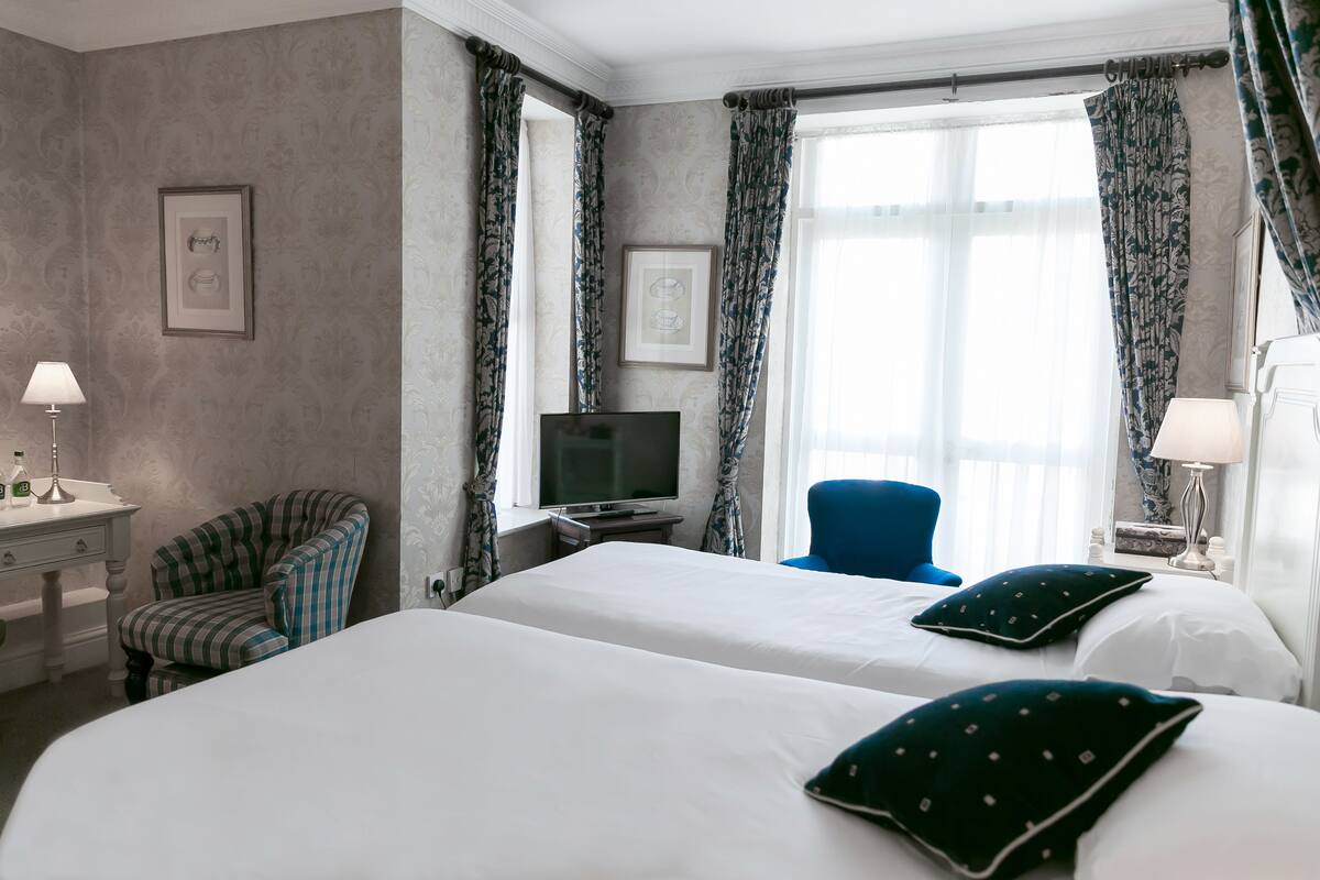 A hotel room with two beds, a TV, a desk with a lamp, a chair, and a window with curtains.
