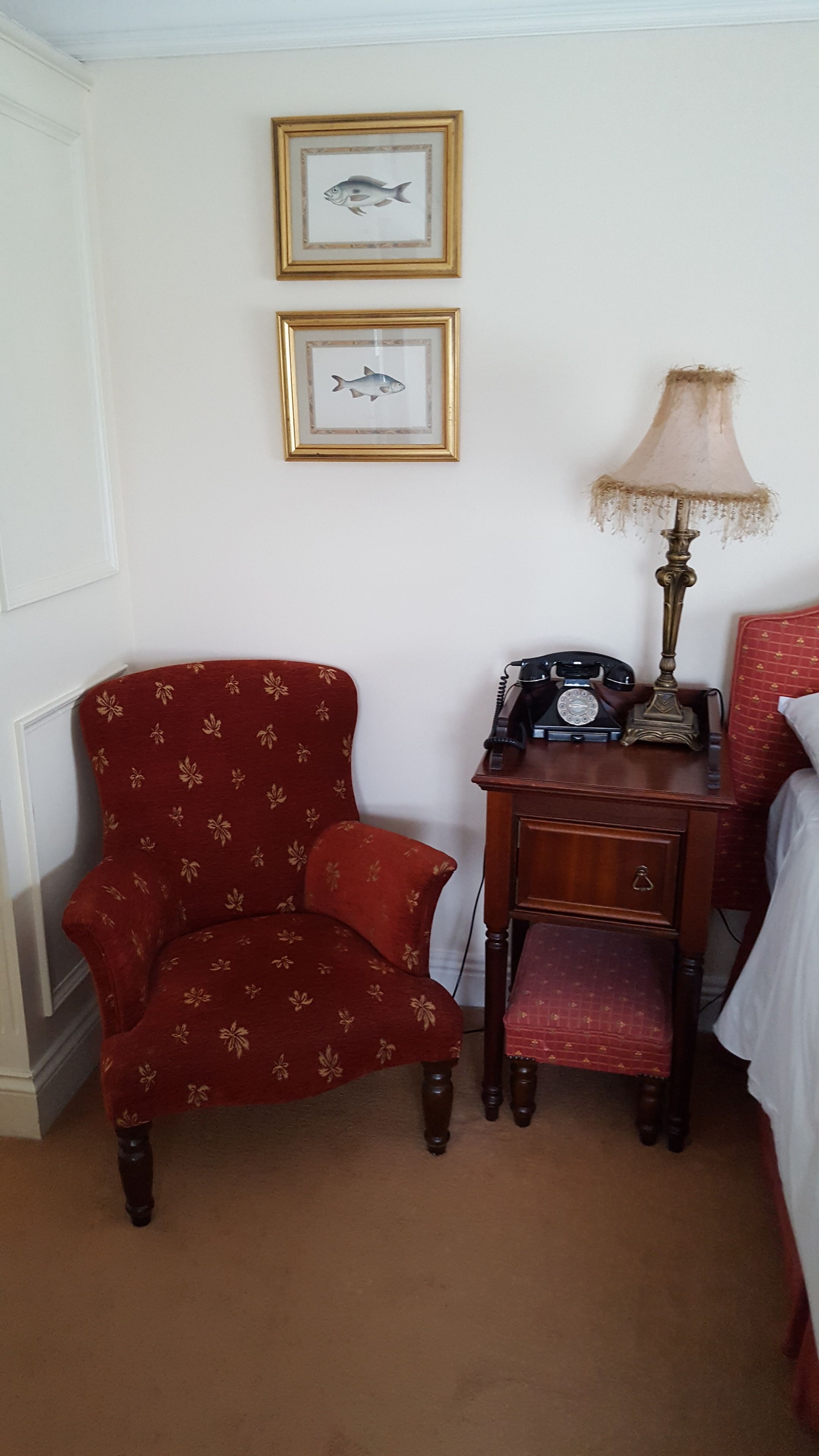 A room with a red armchair, a wooden desk with a telephone and lamp, and two framed fish pictures on the wall.