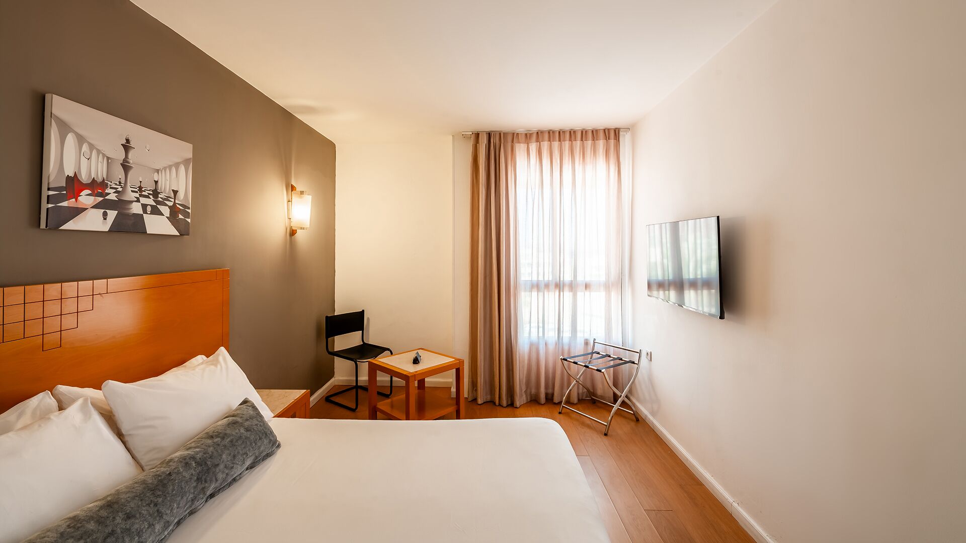 Habitación de hotel con cama, una silla, una mesa, televisor y un cuadro en la pared.