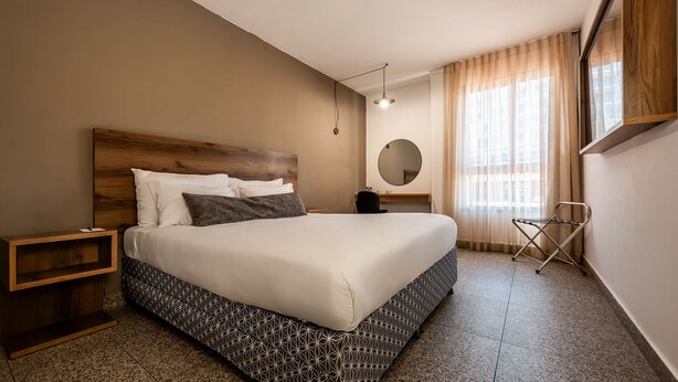 A hotel room with a large bed, a wooden headboard, a bedside table, a mirror, a window with curtains, and a folding chair. - Jacob Eilat (Eilat)