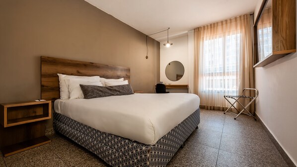 A hotel room with a large bed, a wooden headboard, a bedside table, a mirror, a window with curtains, and a folding chair.