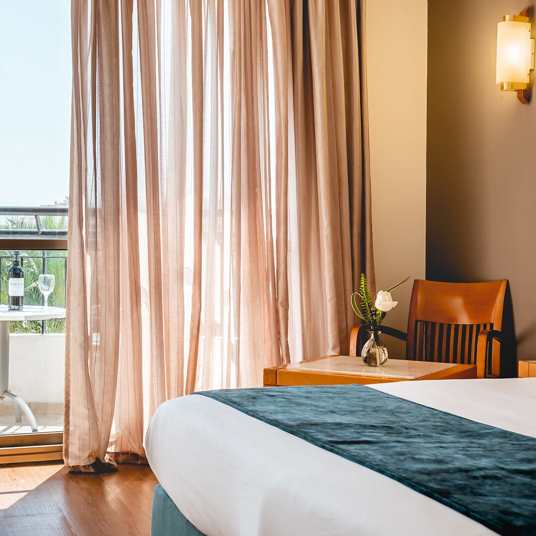 Habitación de hotel con cama, una silla, una mesa con un jarrón de flores y una botella de vino en un balcón.