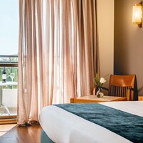 A hotel room with a bed, a chair, a table with a vase of flowers, and a bottle of wine on a balcony.