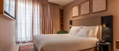 A hotel room with a large bed, a desk with a phone, a chair, and a window with curtains.