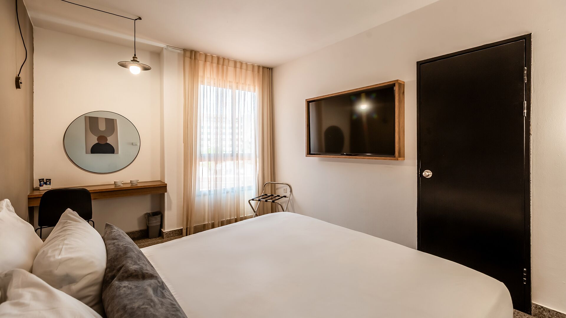 Una habitación de hotel con una cama, un escritorio, una silla, un espejo redondo, un televisor y una puerta.