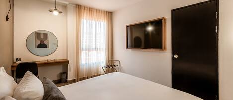 Una habitación de hotel con una cama, un escritorio, una silla, un espejo redondo, un televisor y una puerta.