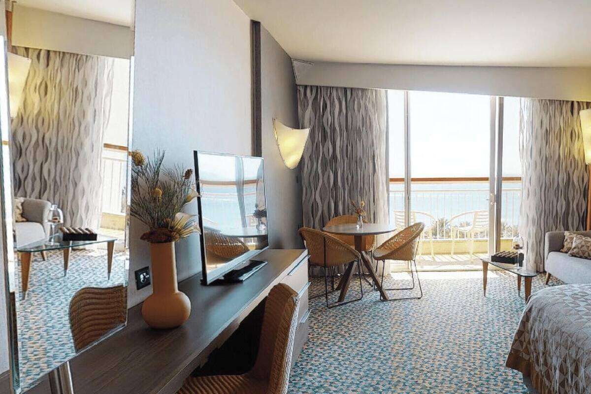 A hotel room with a bed, a desk with a TV, a chair, a sofa, and a balcony with curtains.