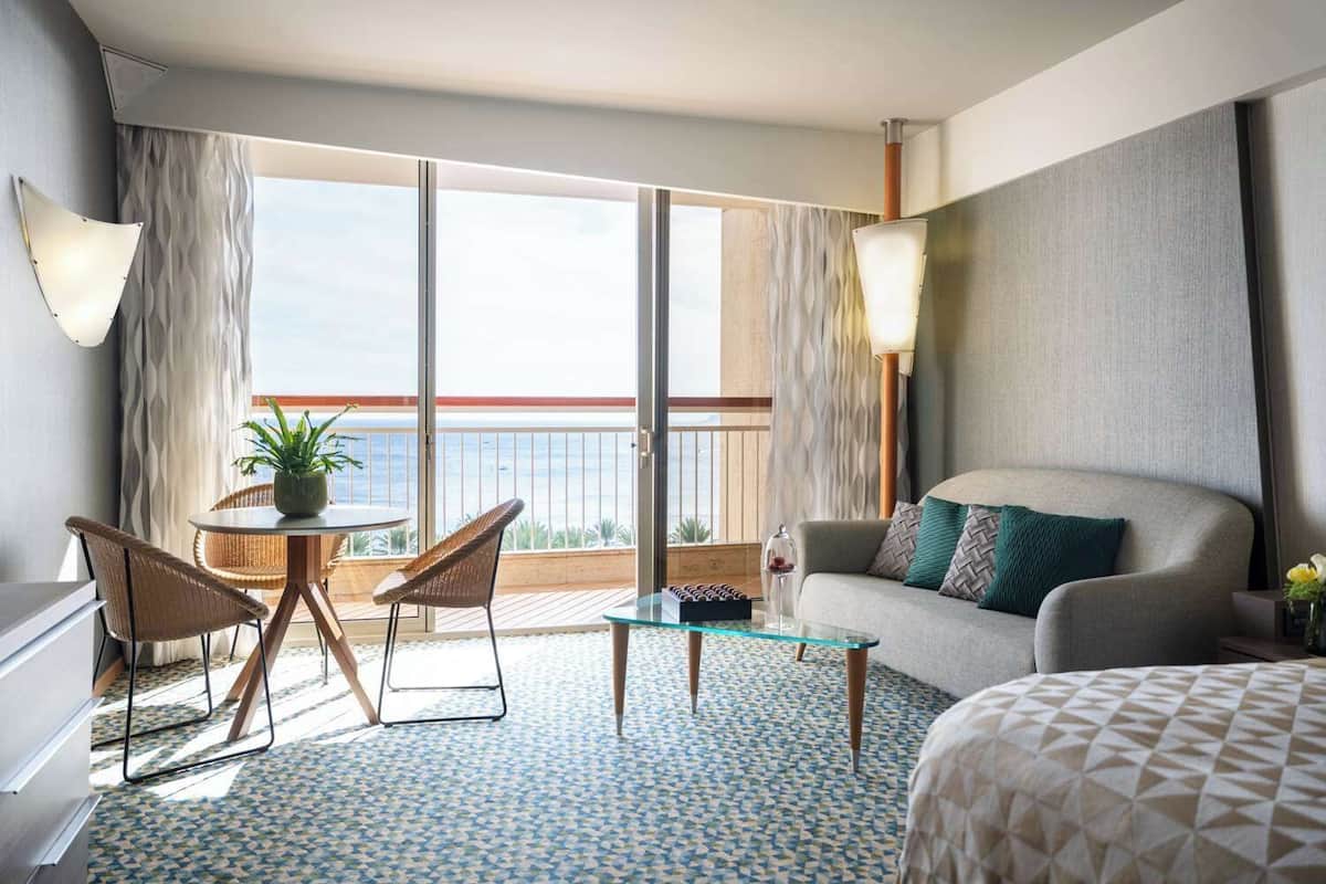 A hotel room with a balcony, a sofa, a dining table with chairs, and a bed.