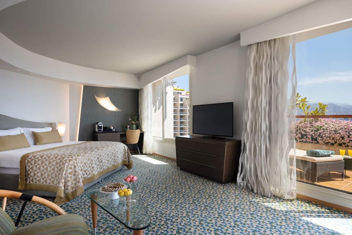 A hotel room with a bed, a TV, a desk, a chair, a glass coffee table, and a balcony with flowers.