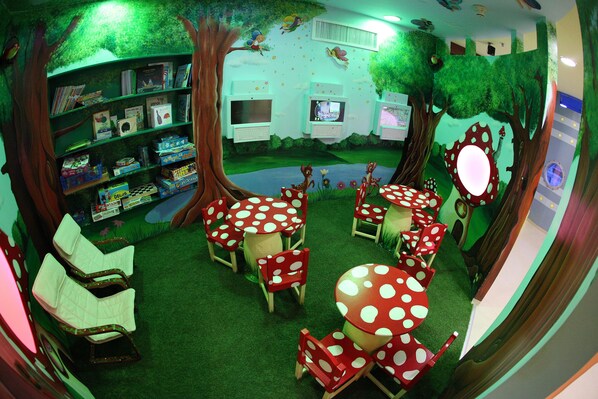 A children's playroom with a forest theme, including a bookshelf, a television, and play tables with chairs.