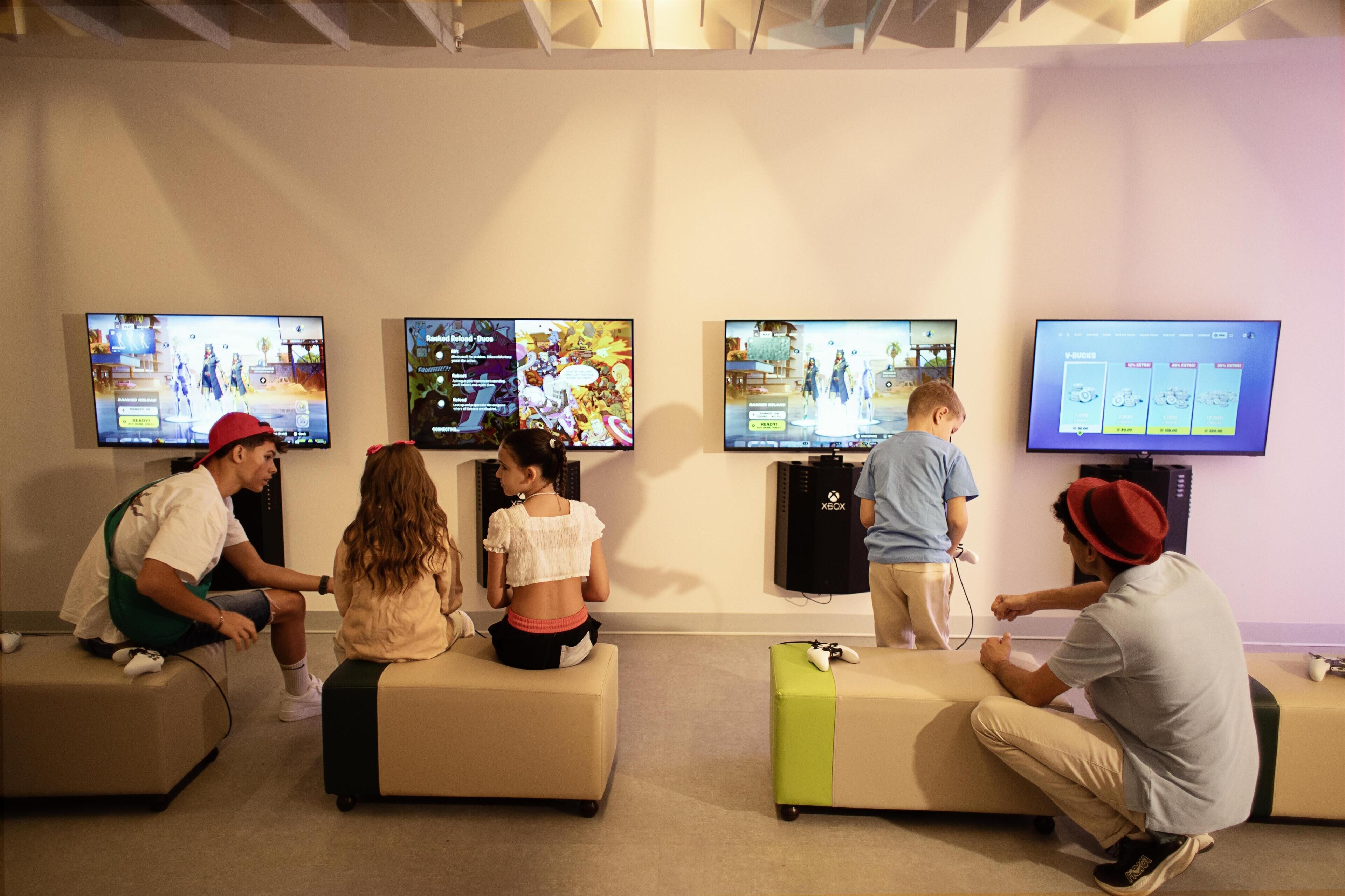 Een game lounge met meerdere schermen waarop videospelletjes worden getoond, zithoeken en Xbox consoles.